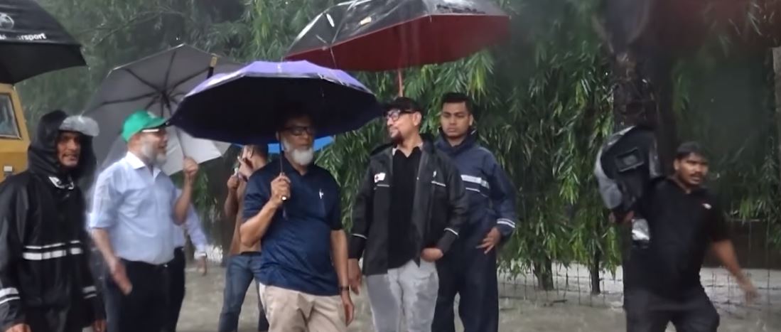 “The State Minister for Railways inspects the submerged railway line while wading through knee-deep water.”
